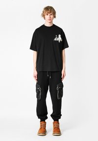 Camiseta negra oversize con un estampado gráfico en el pecho, combinada con pantalones negros ajustados que tienen bolsillos con cremallera y cordón. Botas marrones con cordones.