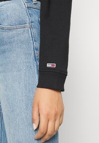 Top negro de manga larga con puños acanalados y un pequeño logotipo bordado. Combinado con jeans de mezclilla azul claro, mostrando detalles de la mano y la manga.
