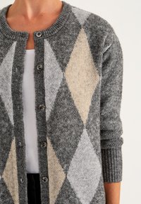 Vrouw in een grijs, beige en lichtgrijs ruitpatroon knopen vest over een witte top met opgerolde mouwen.