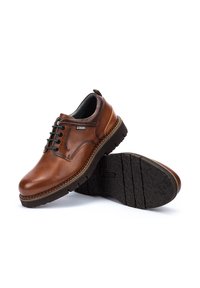 Zapatos de cuero marrón con punta redondeada, diseño de cordones y suela de goma texturizada. Presentan un acento de costura en contraste a lo largo de los bordes.