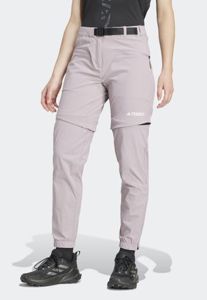 Personne portant un pantalon de randonnée convertible violet clair avec le logo Terrex et une ceinture noire, assorti à des chaussures de sport noires et des chaussettes.