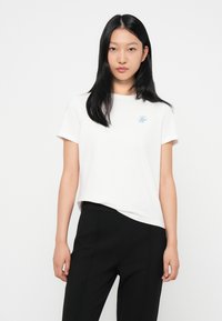 T-shirt bianca in cotone a maniche corte, con logo ricamato blu sul lato sinistro del petto. Abbinata a pantaloni neri.