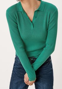 Gestreiftes grünes Poloshirt mit Kragen, zwei Knöpfen im Ausschnitt und langen Ärmeln. Getragen mit dunkelblauen Jeans. Fokus auf der Stofftextur und Farbe.