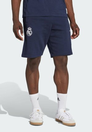 Man draagt marineblauwe Real Madrid-broek, witte Adidas-kortere sokken en witte sneakers met bruine zolen en staat op een lichte achtergrond.