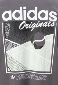 Pelēka T-kreklā ar grafiku. Dizainā ir iekļauta "adidas Originals", tenisa bumba un laukuma elementi. Teksts: "1972. gada TENISA KLUBS."