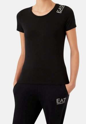 Kobieta ubrana w czarną koszulkę z krótkim rękawem i spodnie, obie z białymi logo EA7 Emporio Armani na ramieniu i udzie, z rękami w kieszeniach.