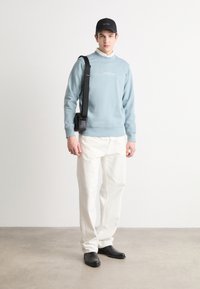 Gaiši zils krekls ar ribinātiem piedurknēm, zem tā balts krekls ar apkakli, krēmkrāsas bikses un melnas kurpes. Krūšu soma pār plecu.