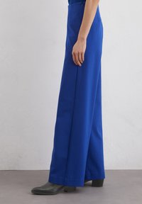 Pantalons larges bleus avec une texture lisse, fermeture éclair latérale et taille haute. Associés à des bottines noires à talons.