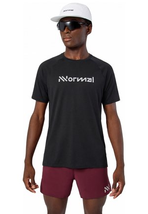 Homme portant un t-shirt noir avec un logo, un short bordeaux, une casquette blanche avec un logo et des lunettes de sport noires, debout devant un fond blanc.