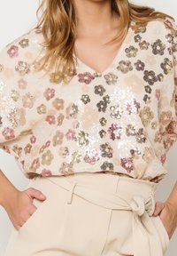 Donna che indossa una blusa floreale beige con paillettes infilata in pantaloni beige a vita alta con cintura annodata, mani in tasca.