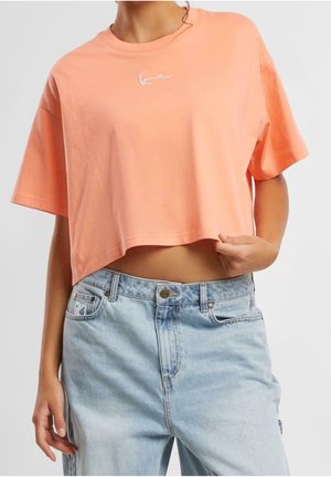 Person iført en ferskenfarvet cropped t-shirt med hvidt logo og lyseblå high-waisted jeans, der holder i t-shirtens kant.
