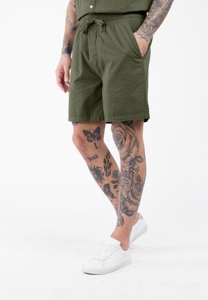 Mann in olivgrünen Shorts und weißen Turnschuhen, dessen tätowierte Beine vor einem weißen Hintergrund sichtbar sind.
