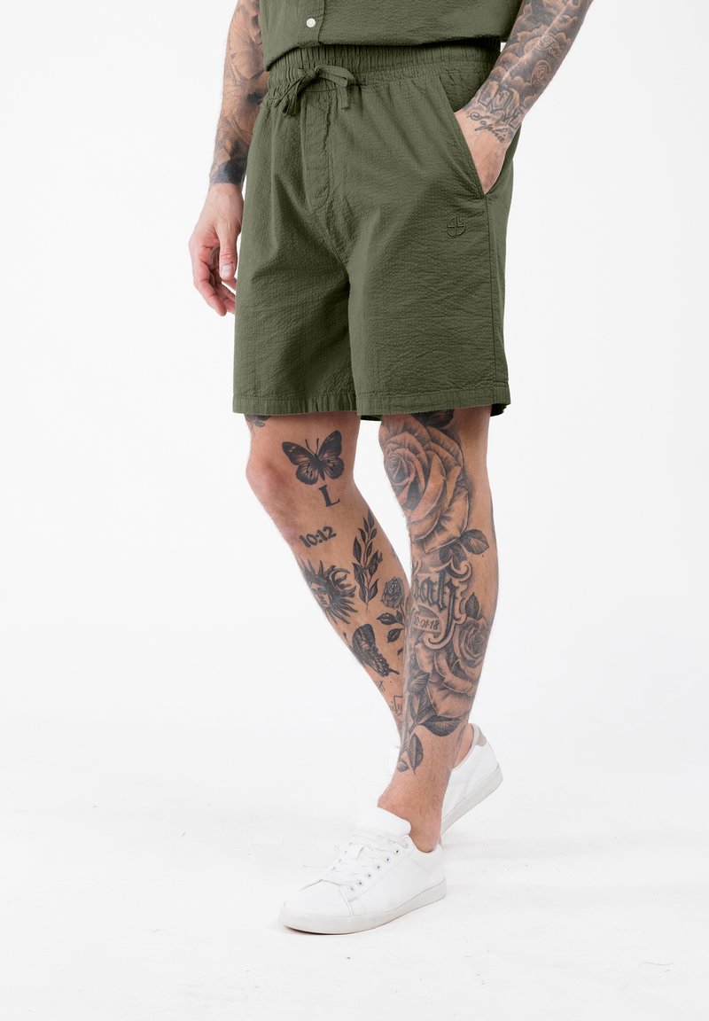 Uomo che indossa pantaloni corti verde oliva e sneaker bianchi, con le gambe tatuate in mostra su uno sfondo bianco.