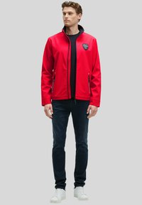 Giacca softshell rossa con colletto alto, tasche con zip e dettaglio del logo sul petto, abbinata a jeans scuri e sneakers bianche.