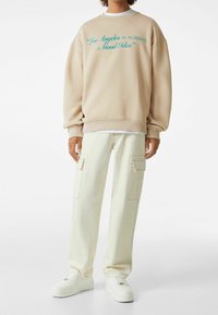 Persona che indossa una felpa beige con testo turchese "Los Angeles è sempre una buona idea", pantaloni cargo crema e sneakers bianche su sfondo bianco.