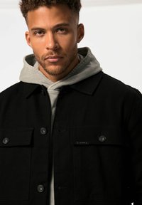 Zwarte button-up jas met twee borstzakken, gemaakt van een dikke stof. Een grijze hoodie is eronder gelaagd, waarbij de capuchon zichtbaar is.