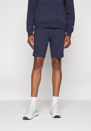 Mand iført marineblå shorts og matchende hættetrøje, hvide sokker og hvide sneakers, stående mod en ensfarvet lys baggrund.