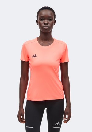 Femme portant un t-shirt de sport corail à manches courtes et un legging noir, tous deux avec le logo Adidas, debout devant un fond uni.