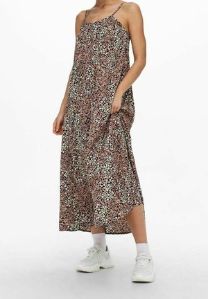 Kobieta ubrana w sukienkę maxi bez rękawów w kwiatowy wzór z cienkimi ramiączkami oraz białe trampki z skarpetkami, trzymająca bok sukienki.