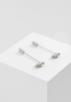 Paar zilverkleurige metalen oorsteker met bolvormige uiteinden, tentoongesteld op een wit kubusvoetstuk tegen een grijze achtergrond.
