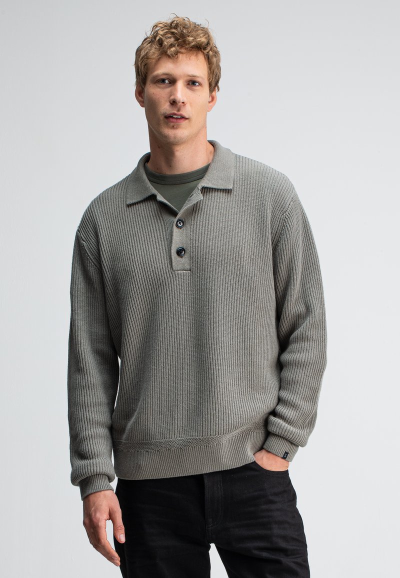 Lichtgrijze geribde pullovertrui met een kraag, drie zwarte knopen en een gestructureerde stof; gedragen over een donkergroen shirt en zwarte jeans.