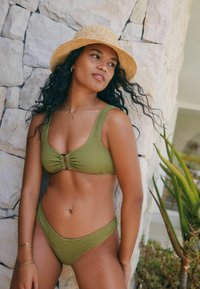 Olivgrünes Bikini-Set aus strukturiertem Stoff, mit breiten Trägern und einer Knoten-Detail vorne. Gestylt mit einem Strohhut vor einer Steinwand.