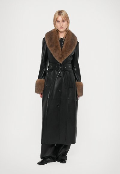 Woman wearing a long black leather coat with brown fur collar and cuffs, standing against a plain white background.