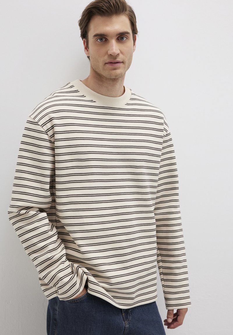 Beige Langarmshirt mit feinen dunklen Streifen, rundem Ausschnitt, lässiger Passform, weichem Stoff, kontrastierenden Manschetten, kombiniert mit dunkelblauen Jeans.