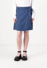 Denim omslagskjørt i blått med knytelukking i livet, med skrå sømmer og en lett utsvinget kant. Kombinert med svarte platåsko.