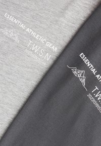 Két összehajtott póló, az egyik világosszürke, a másik sötétszürke, mindkettőn fehér szöveg: „ESSENTIAL ATHLETIC GEAR T.W.S.N” és hegyvonal rajz.
