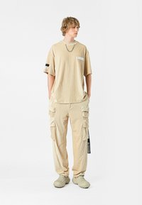 Camisa de manga corta de rayas en color beige con bolsillo en el pecho y detalle de cadena, acompañada de pantalones cargo a juego que cuentan con múltiples bolsillos y etiquetas.