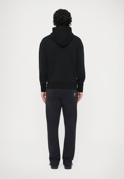 Uomo con capelli ricci che indossa un felpa nera con cappuccio e pantaloni della tuta neri con un logo sulla tasca posteriore, in piedi di fronte a un muro bianco.