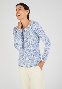 Maglietta a maniche lunghe di colore azzurro chiaro con un motivo floreale in blu scuro e bianco. Texture liscia con scollatura rotonda, abbinata a pantaloni crema.