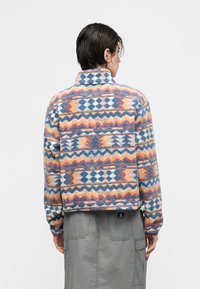 Άτομο με κοντά σκοτεινά μαλλιά φοράει πολύχρωμο φλις ζακέτα με σχέδια και γκρι φούστα cargo, κοιτώντας προς τα πίσω σε απλό φόντο.