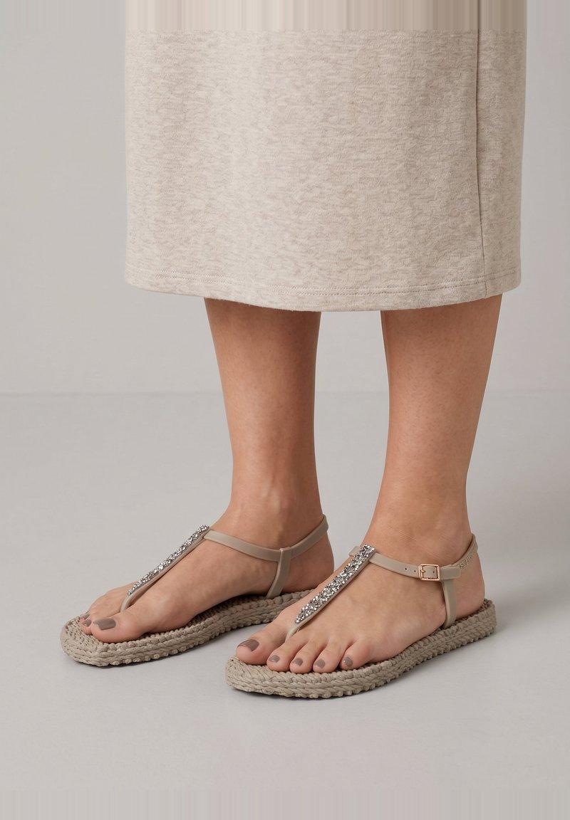 Voeten met beige platte sandalen met sierlijke bandjes en gespsluitingen, gecombineerd met een lichtbeige knie-lange rok.