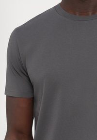 Gros plan d'une personne portant un t-shirt gris foncé uni à manches courtes et col rond sur un fond neutre.