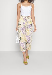 Žena ve bílém krátkém topu, květovaných vysokých kalhotách s širokými nohavicemi, zelených otevřených lodičkách a nese malou bílou kabelku.