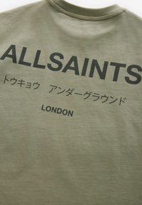 Olive spalvos medvilninis marškinėlis su apvaliu kaklu, ant kurio yra ryškus juodas užrašas "ALLSAINTS" ir japonų simboliai, o žemiau - "LONDON".