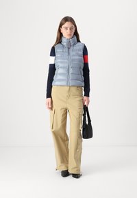 Gilet rembourré bleu clair avec col haut, en tissu léger ; associé à un pantalon cargo kaki avec des poches latérales et un sac à main noir.