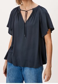 Blouse bleu marine foncé avec un col en V, des manches froncées et un détail à nouer au niveau du col. Tissu doux et léger avec une texture subtile.