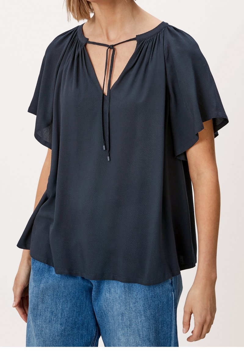 Blouse bleu marine foncé avec un col en V, des manches froncées et un détail à nouer au niveau du col. Tissu doux et léger avec une texture subtile.