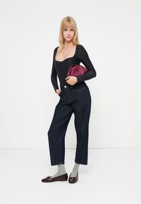 Body noir à manches longues, jeans taille haute bleu marine, chaussettes grises et mocassins bordeaux, tenant une pochette bordeaux.