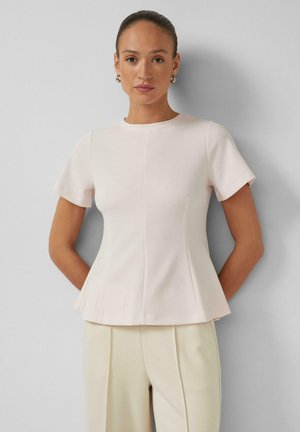 Vrouw met naar achteren gebonden haar, zilverkleurige oorringen, een lichtroze nauwsluitend shirt met korte mouwen en een uitlopende zoom, en crème kleurige broek.