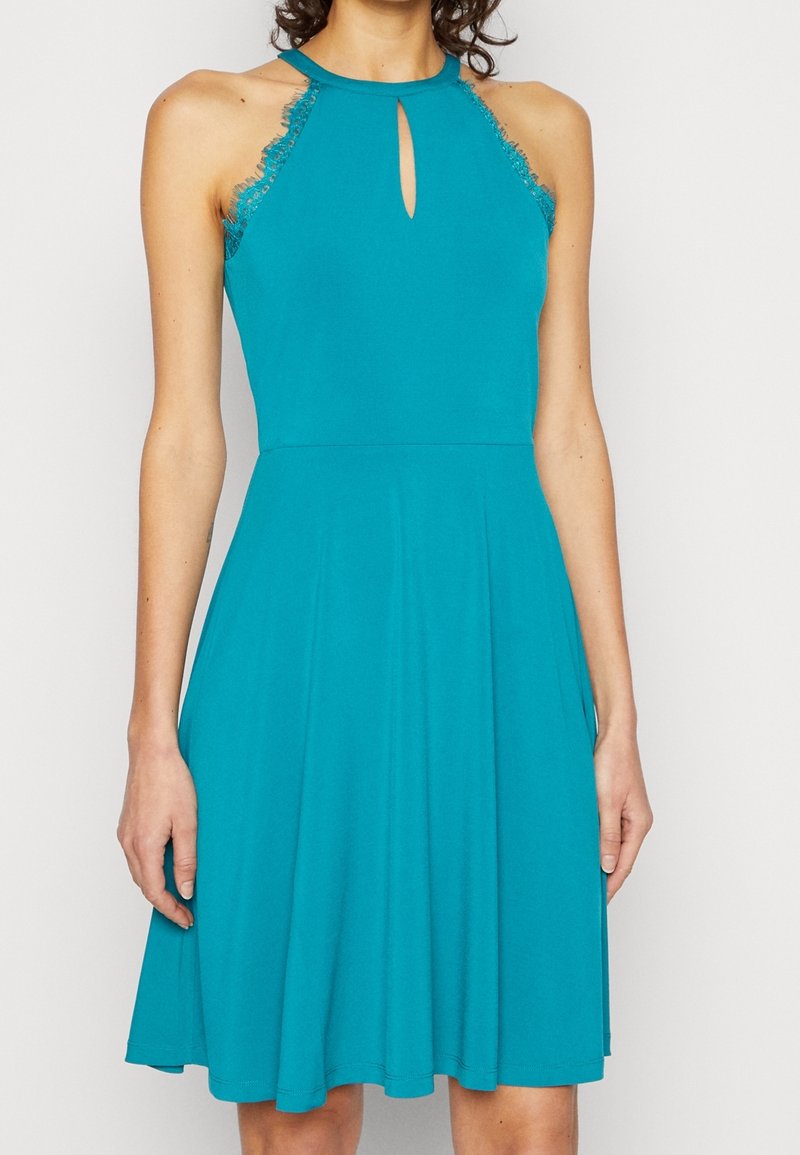 Femme portant une robe sans manches bleu sarcelle avec une bordure en dentelle et un décolleté en trou de serrure à l'avant, debout devant un fond uni.