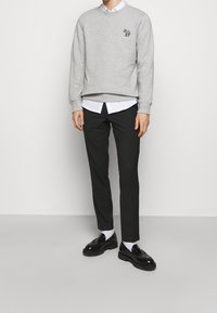 Sudadera gris con un pequeño diseño bordado, superpuesta a una camisa blanca con cuello. Combinada con pantalones negros y mocasines negros.