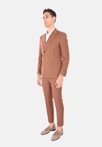 Completo marrone a doppio petto con dettagli in oro, vestibilità sartoriale e pantaloni slim, abbinato a scarpe di colore chiaro. Texture liscia.