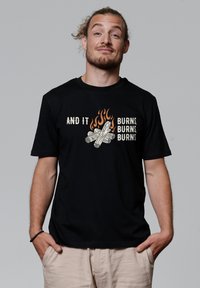 Zwarte katoenen t-shirt met geprinte tekst en een grafiek van een kampvuur met houtblokken en vlammen in oranje en crèmekleuren. Korte mouwen en ronde hals.