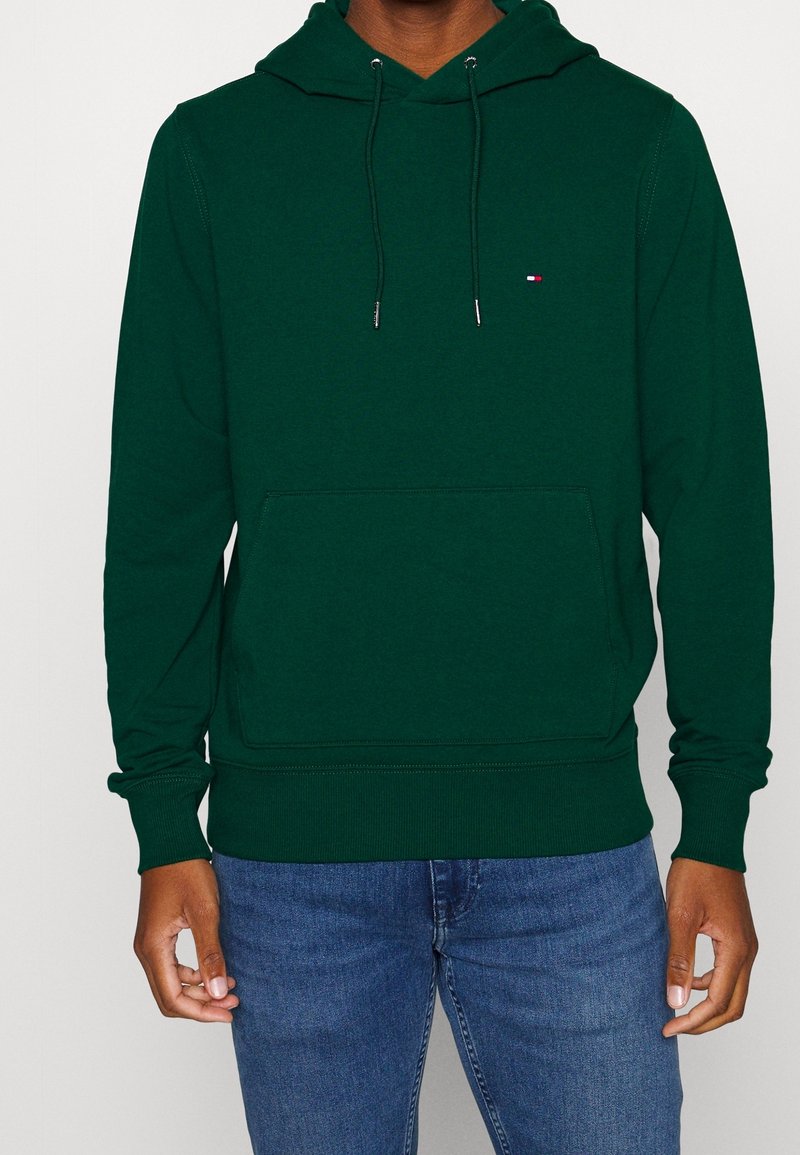 Homme portant un sweat à capuche vert foncé avec cordons et poche avant, associé à un jean bleu, debout devant un fond uni.