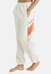 Krämfärgade sweatpants med röda sömmar och orange detaljer; elastisk midja och sidofickor. Vänster ben har "ROXY"-logotypen.