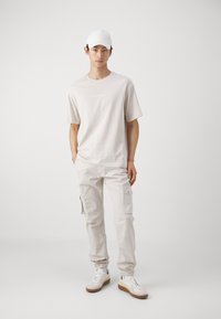 Camiseta beige clara con logo, combinada con pantalones cargo claros que tienen bolsillos laterales y puños elásticos. Calzado: zapatillas blancas con acentos en beige.